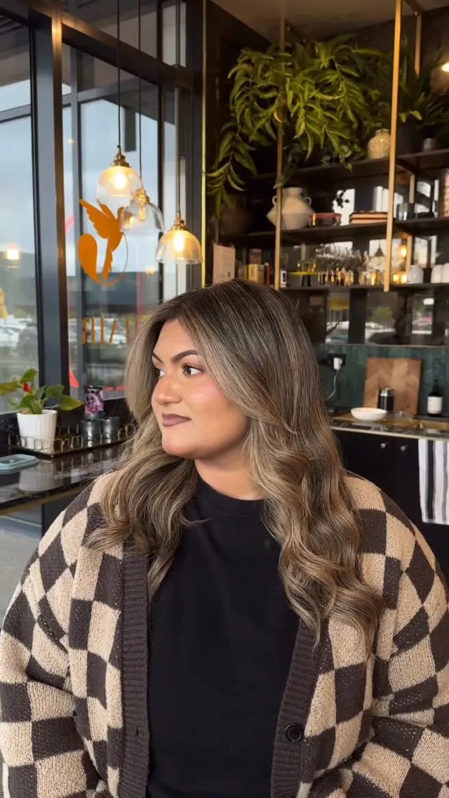 A client with long honey-blonde waves and blended dark roots, smiling in the salon